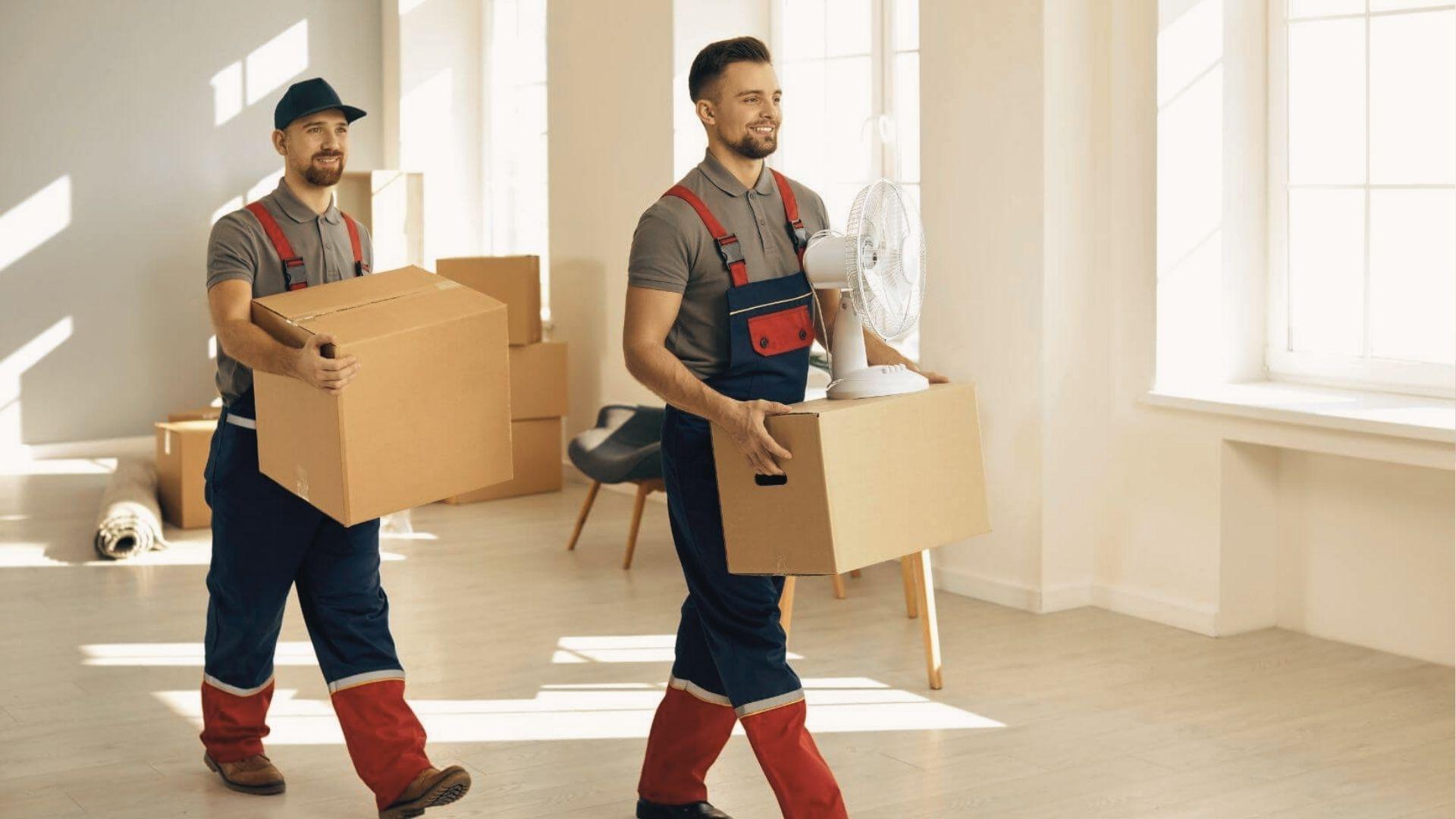 Movers in red-and-blue overalls carry boxes and a fan into a bright, empty apartment.