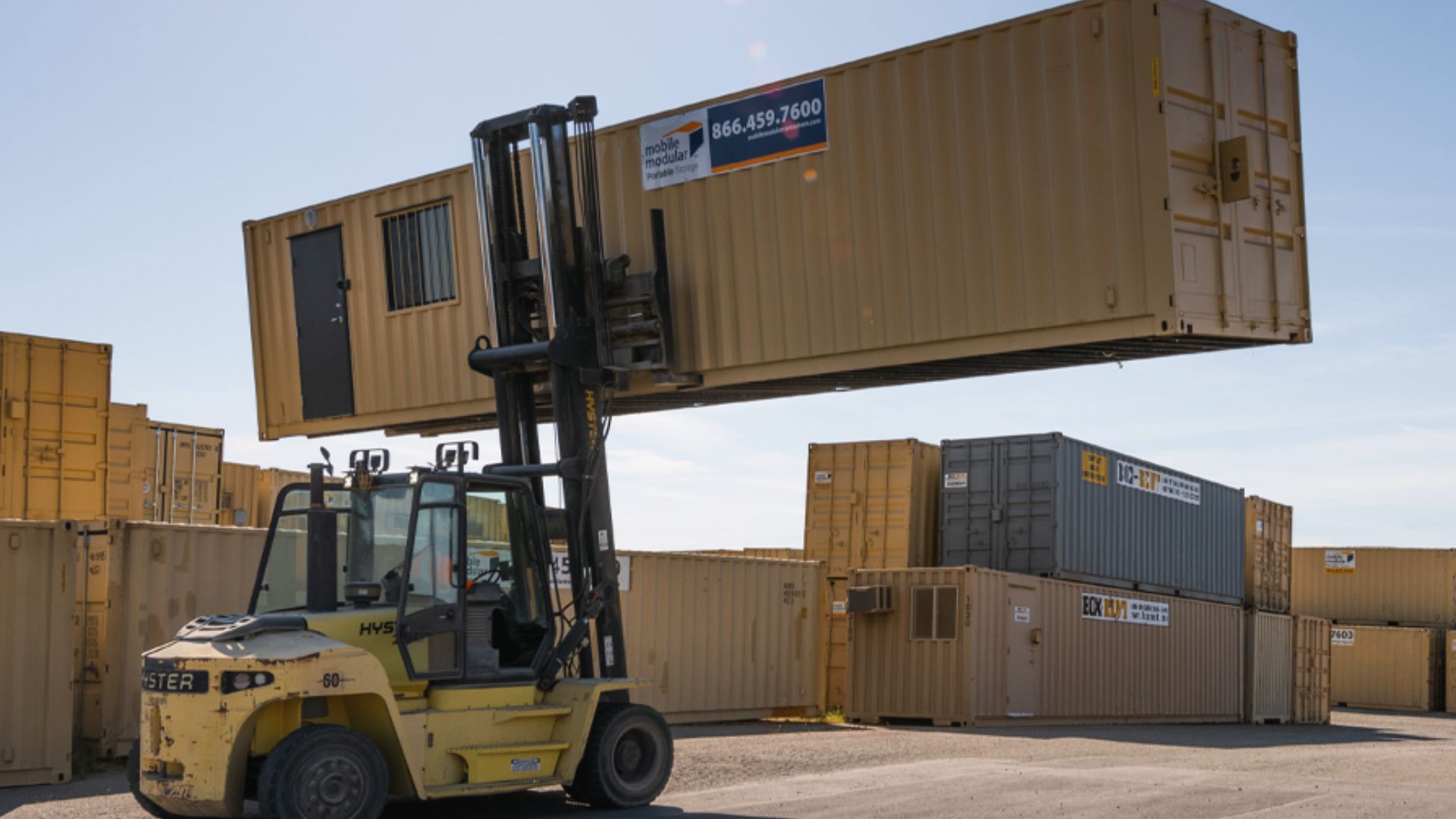 Yellow forklift lifting a tan modified shipping container with a door and window in a storage yard.