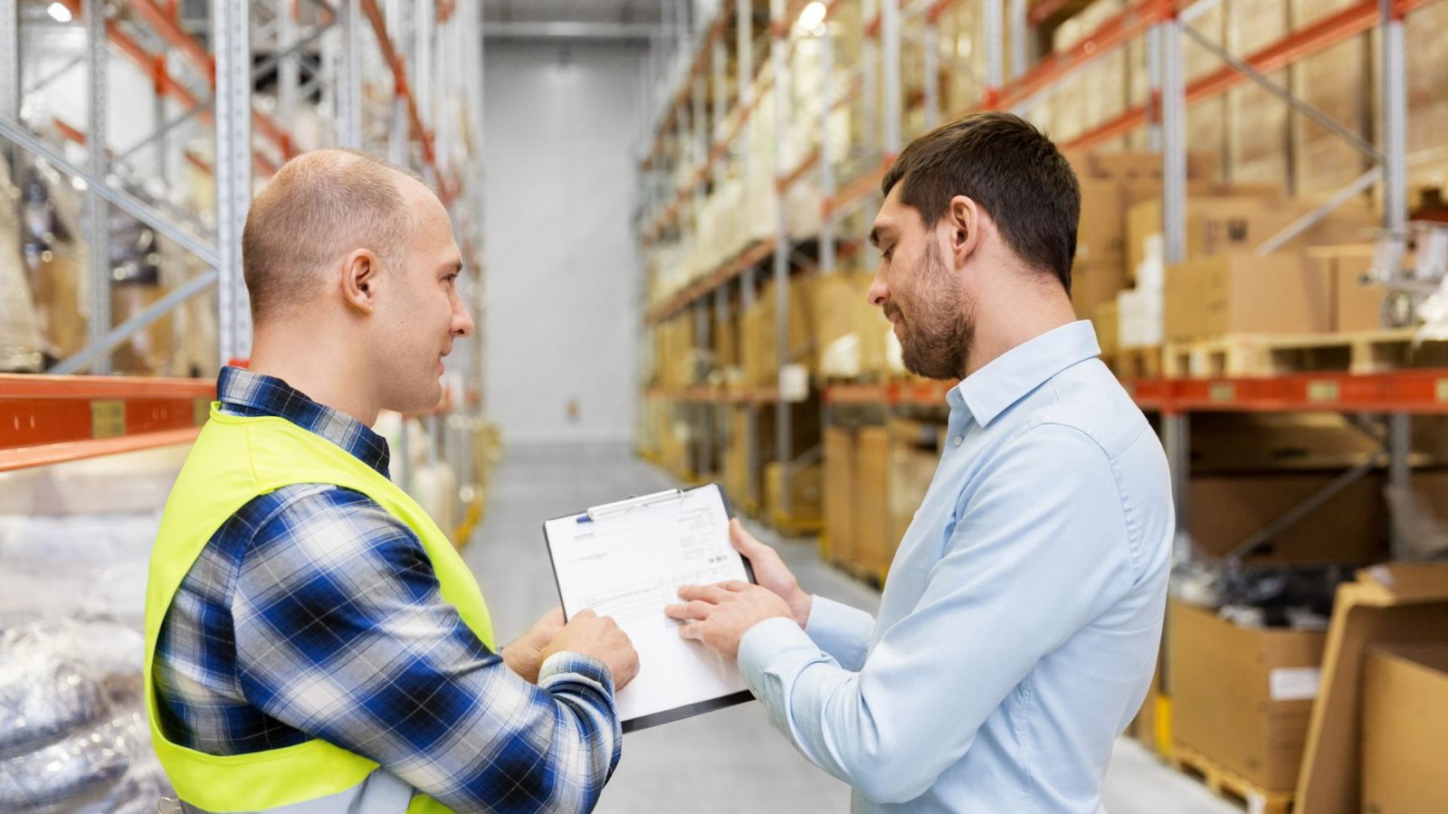 Two warehouse staff members reviewing a shipping manifest on a clipboard between storage racks.