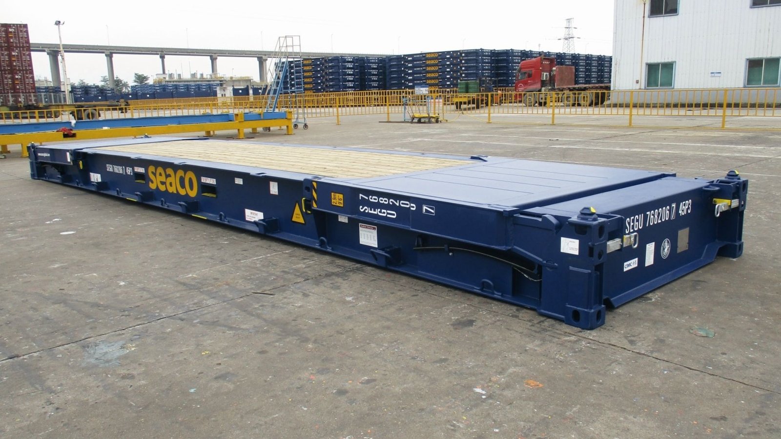 Dark blue flat rack container without sides or roof, branded “seaco” on a concrete yard.