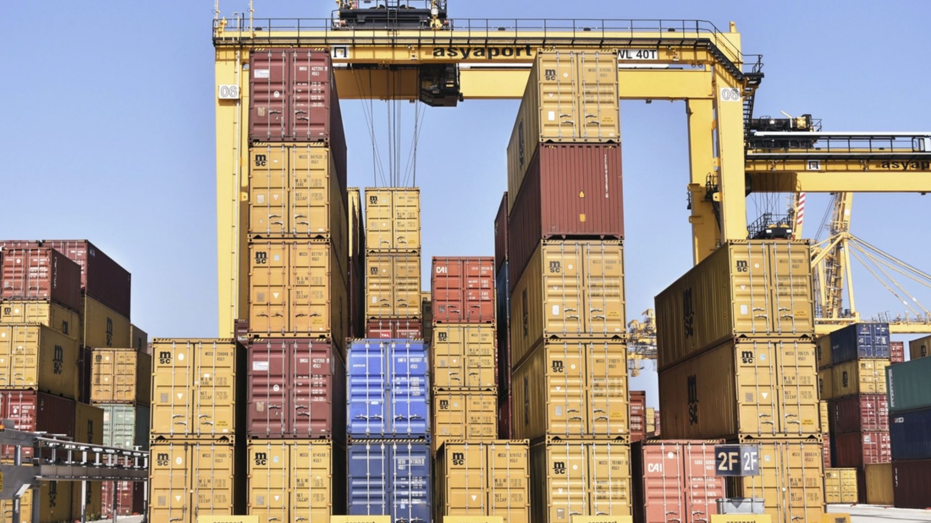 Yellow gantry crane lifting multiple stacked yellow and red shipping containers at a port terminal.