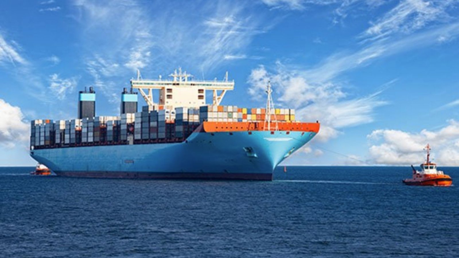A large blue container ship loaded with colorful cargo sailing on the open ocean with tugboats.