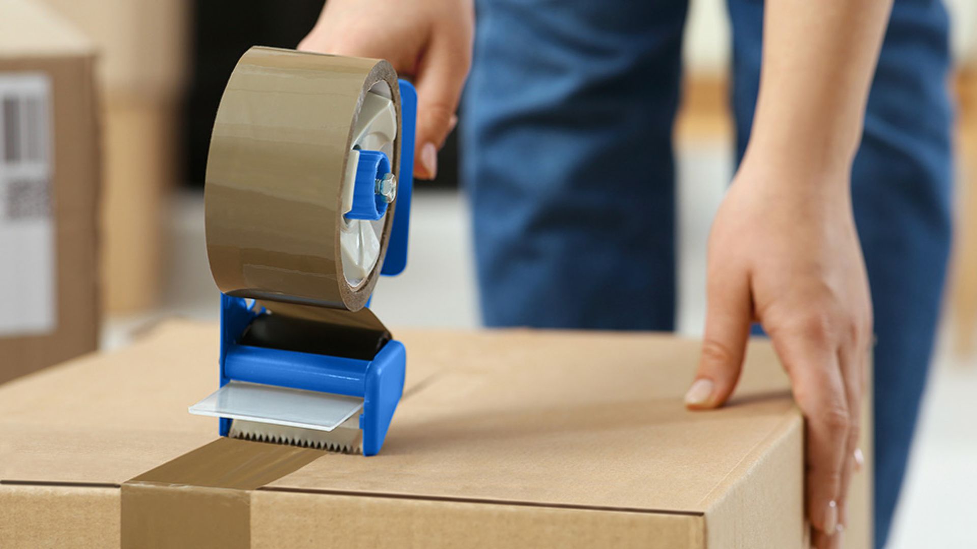 A person operates a tape dispenser to pack the  box, illustrating the importance of proper tape selection for packing.
