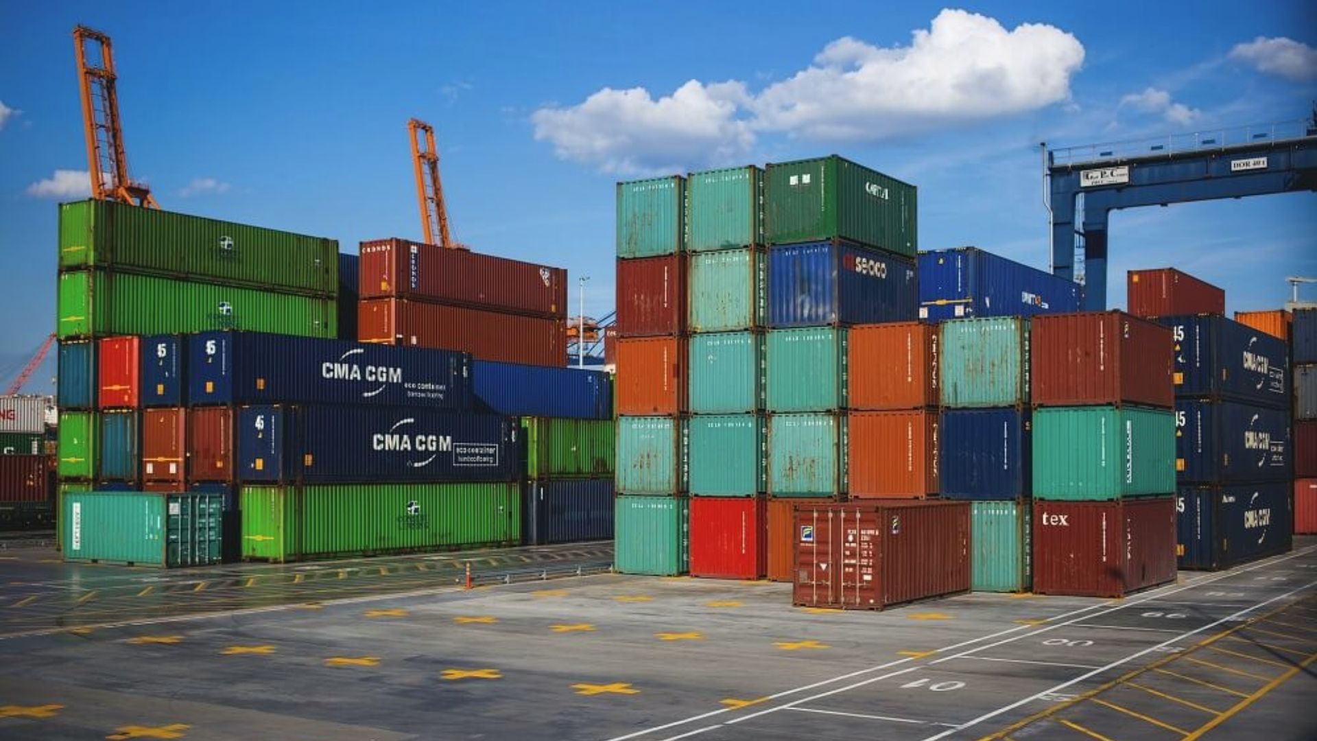 Stacks of green, blue, and red shipping containers at a busy port under a clear sky with cranes in the background.