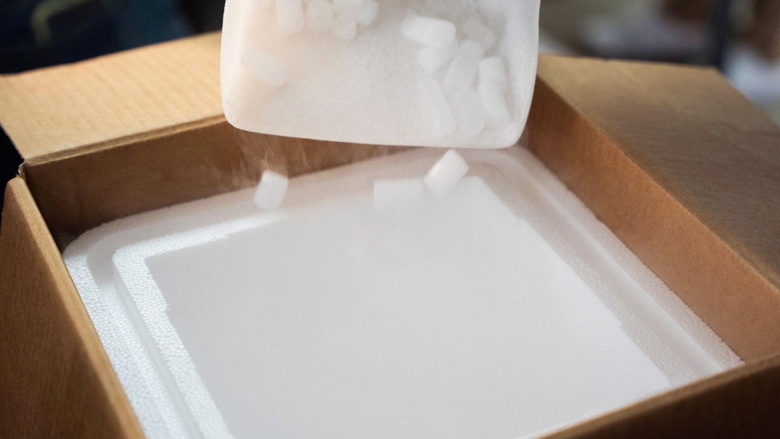 Dry ice pellets being poured into an insulated cardboard shipping box lined with styrofoam.