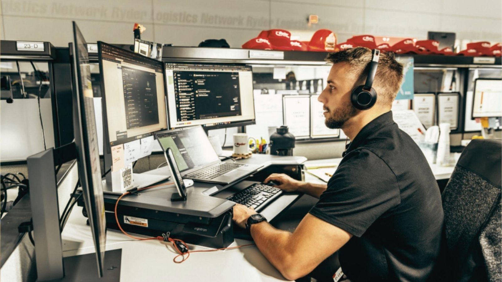 A logistics broker wearing a headset manages freight bookings across multiple monitors and a laptop.